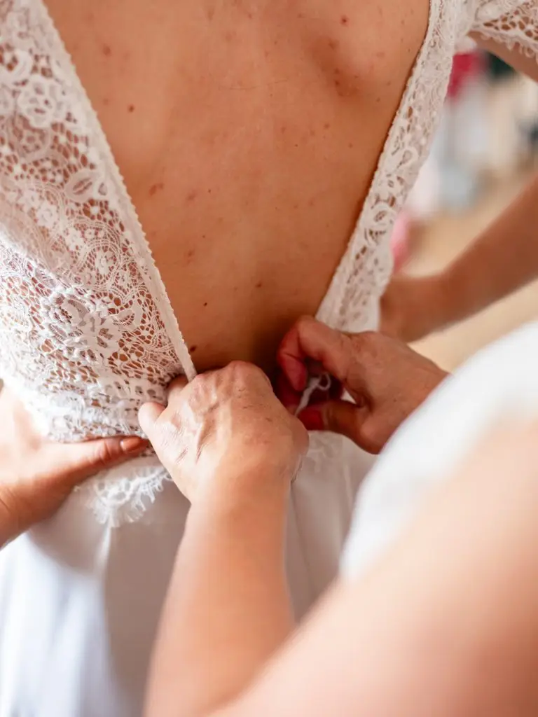 La maman de la mariée boutonne la robe blanche de sa fille.
