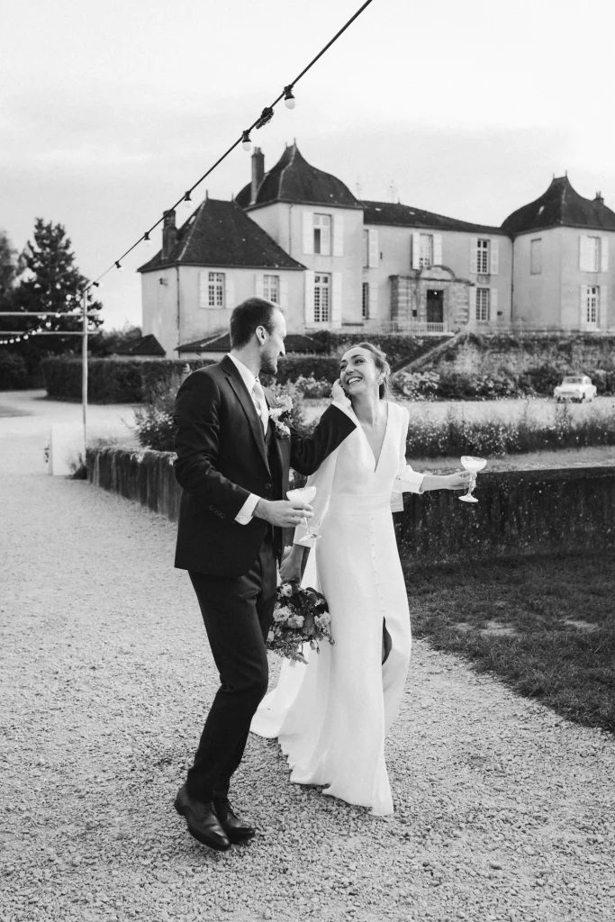 Le marié caresse la joue de sa femme alors qu'ils reviennent de leur séance couple, verres à la main, sous les applaudissements de leurs invités.