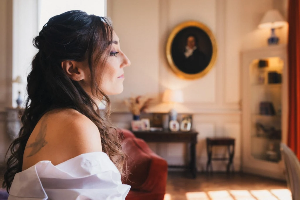 La mariée a l'air pensif, pendant ses préparatifs de coiffure et maquillage ; sa chemise dénude légèrement son épaule et révèle un oiseau tatoué dessus.