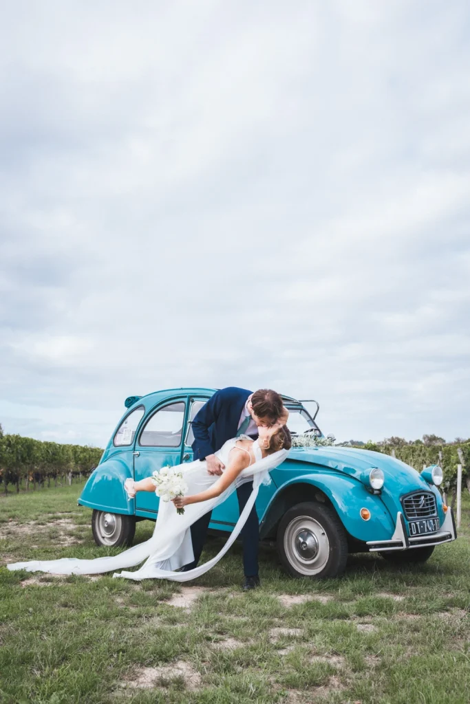 Le marié renverse sa promise pour l'embrasser devant leur voiture de collection bleue, au milieu des vignes, lors de leur mariage en Gironde.