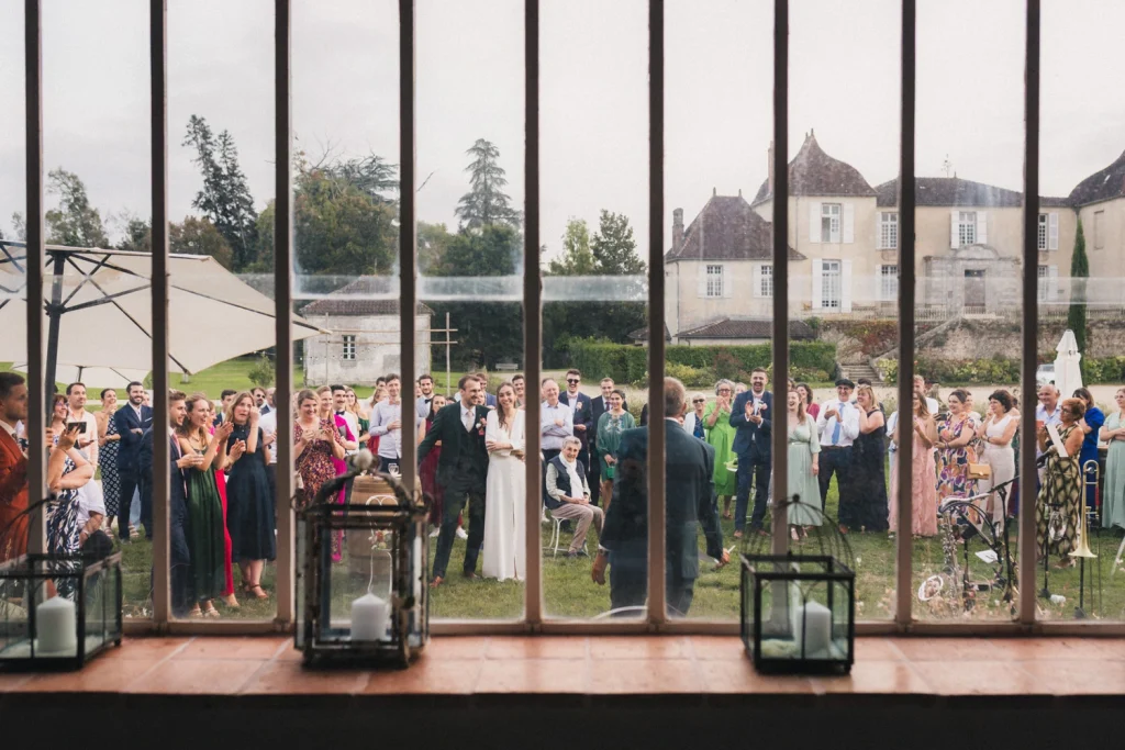Depuis l'intérieur de la salle de réception, on aperçoit les mariés, entourés de leurs invités, qui écoutent attentivement le discours des parents à l'extérieur, lors d'un mariage dans les Landes.