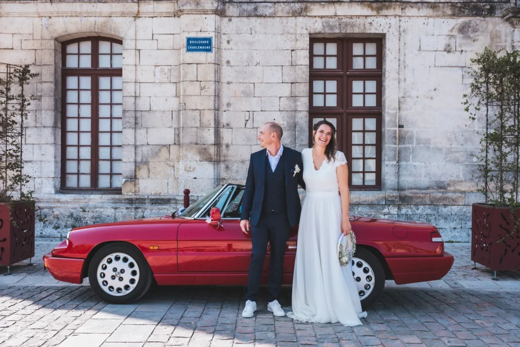 Les mariés prennent la pose devant leur voiture de collection rouge après leur cérémonie civile de mariage en Dordogne.