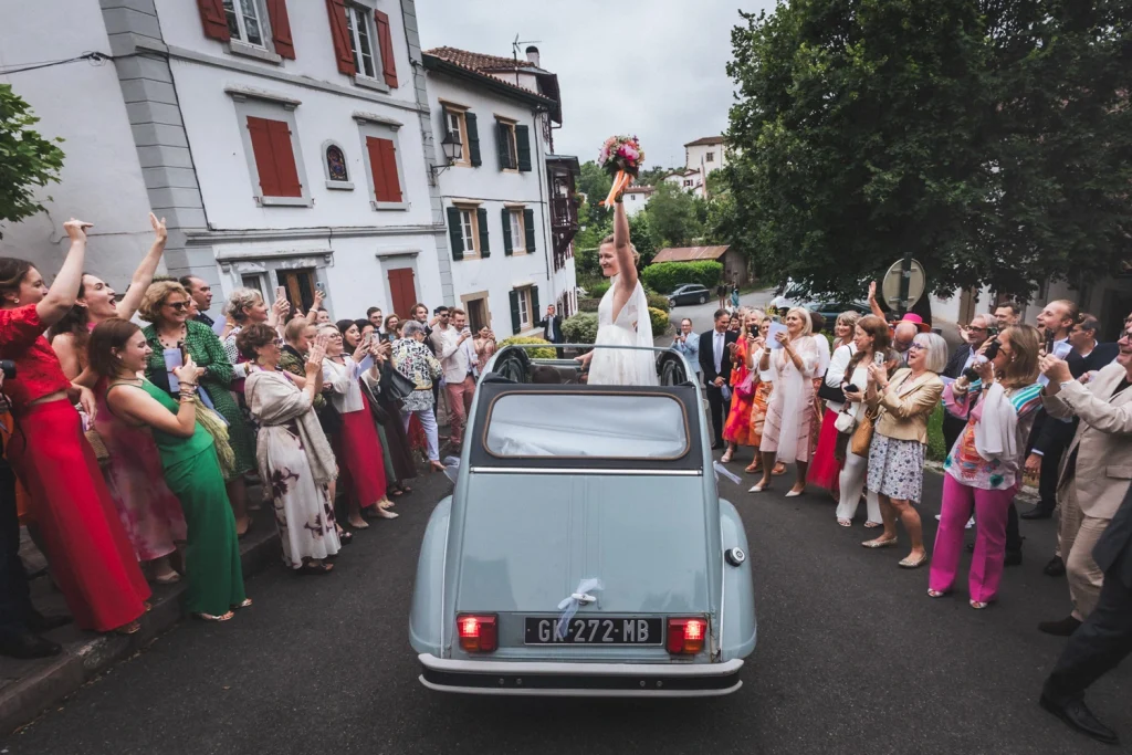 Mariage à Espelette au Pays Basque, où les mariés quittent l'église dans une voiture bleue, la mariée debout sur le siège qui salue une foule d'invités en liesse.