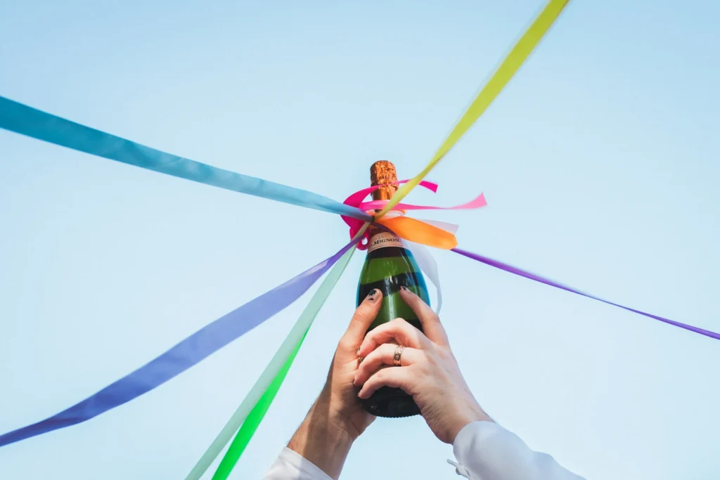 Détail de la bouteille de champagne tenue par les mains des mariés et à laquelle sont accrochés des rubans de couleur, lors d'un mariage à Bordeaux