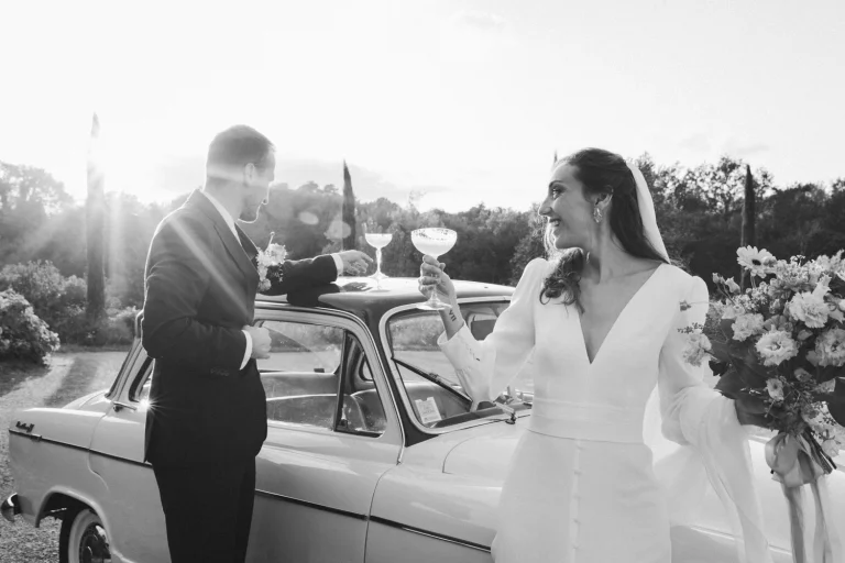 Durant leur séance couple, les mariés trinquent avec leurs verres de cocktail, devant une voiture de collection, dans les derniers rayons du soleil dans les Landes