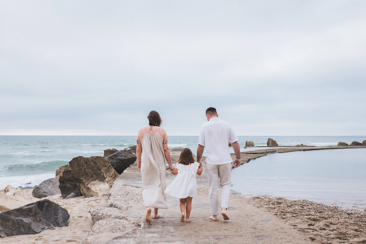 Lors d'une séance famille sur la plage en Gironde, les parents, de dos, tiennent leur fille par la main et s'avancent vers l'océan.