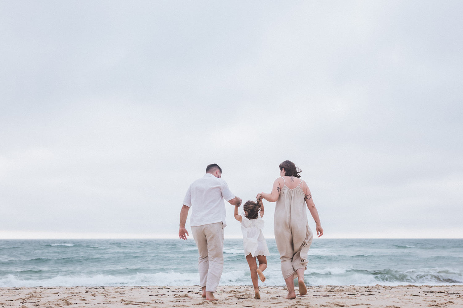 De dos, face à la mer, les parents tiennent chacun par la main leur fille et courent tous ensemble vers l'eau
