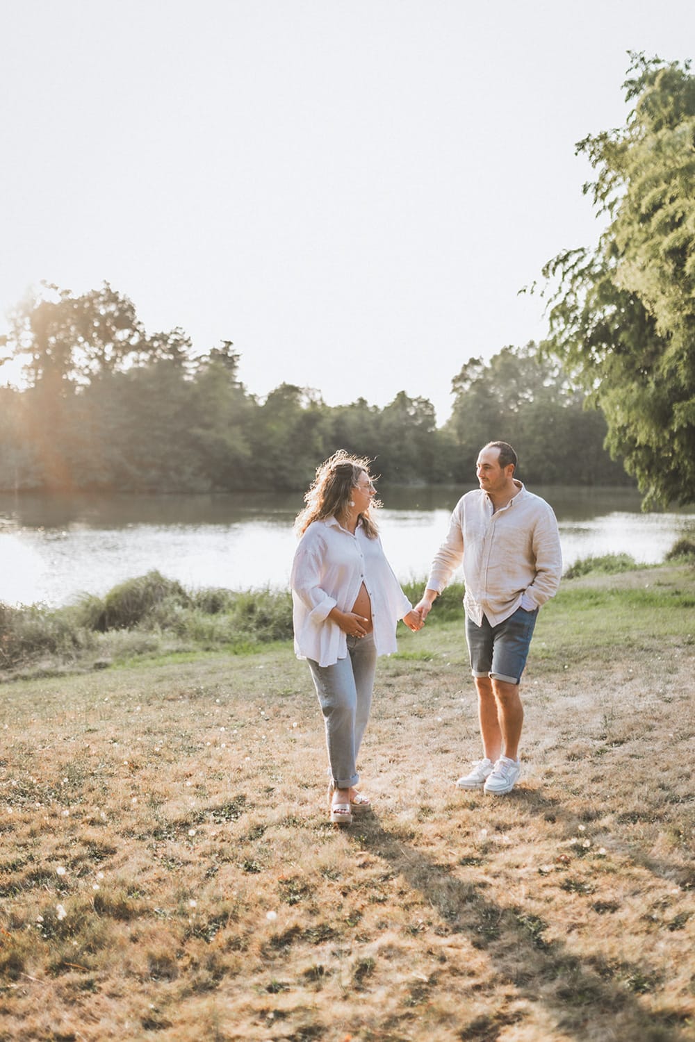 Les futurs parents marchent main dans la main en se regardant au bord d'un lac, une main sur le ventre rond de la femme enceinte, immortalisé par une photographe famille landes