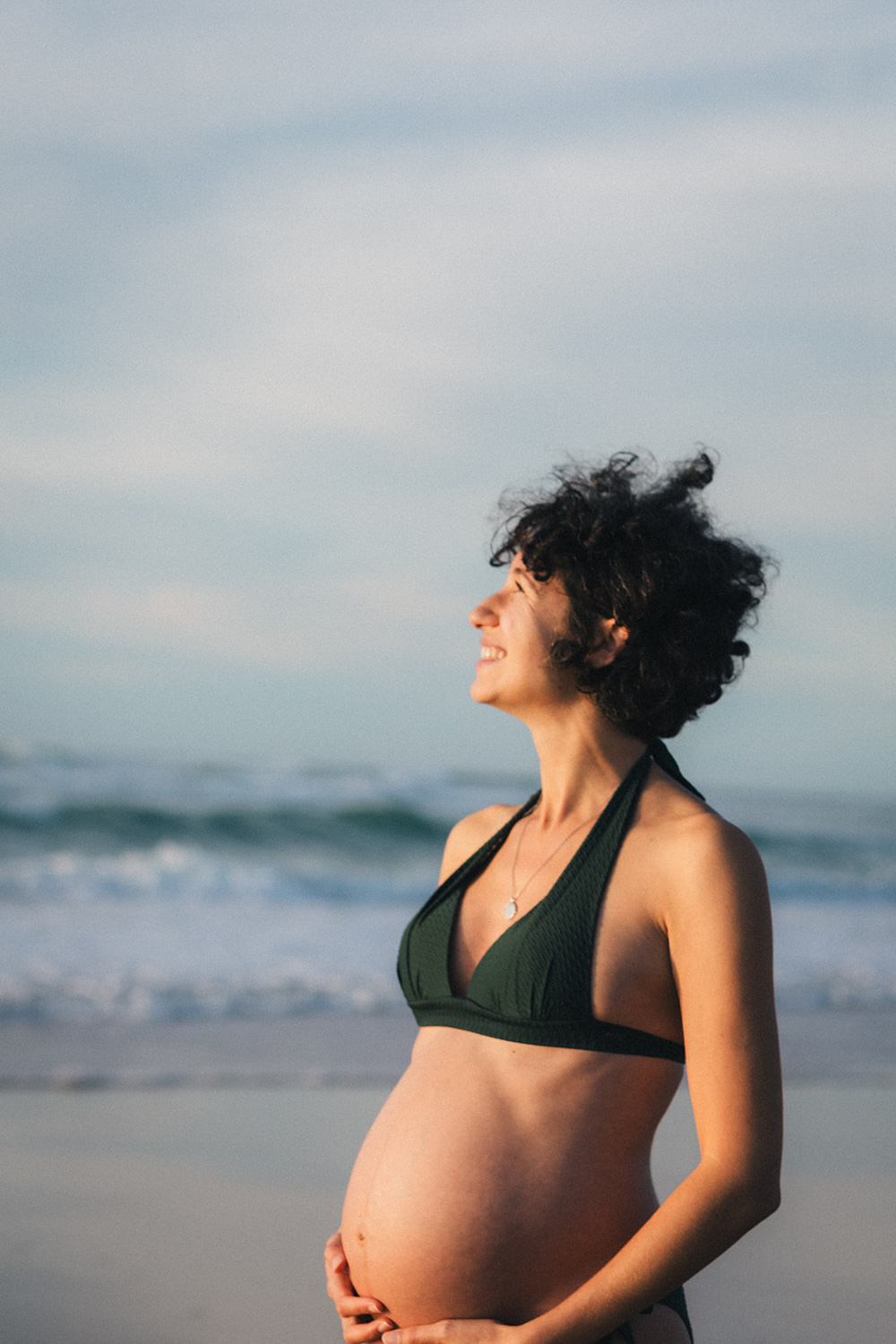 Une future maman en tenue de plage regarde vers le ciel et l'océan, lors d'une séance grossesse réalisée par une photographe famille landes