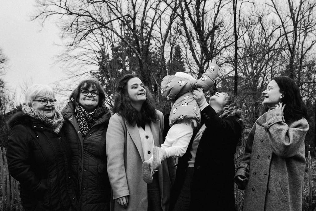 Lors d'une séance famille intergénérationnelle entre femmes, une tante soulève sa petite nièce dans ses bras pendant que les autres la regardent faire.
