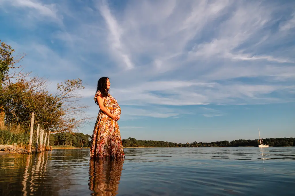 La future maman regarde vers l'horizon, au milieu d'un lac en tenant paisiblement son ventre, avec une photographe grossesse bordeaux