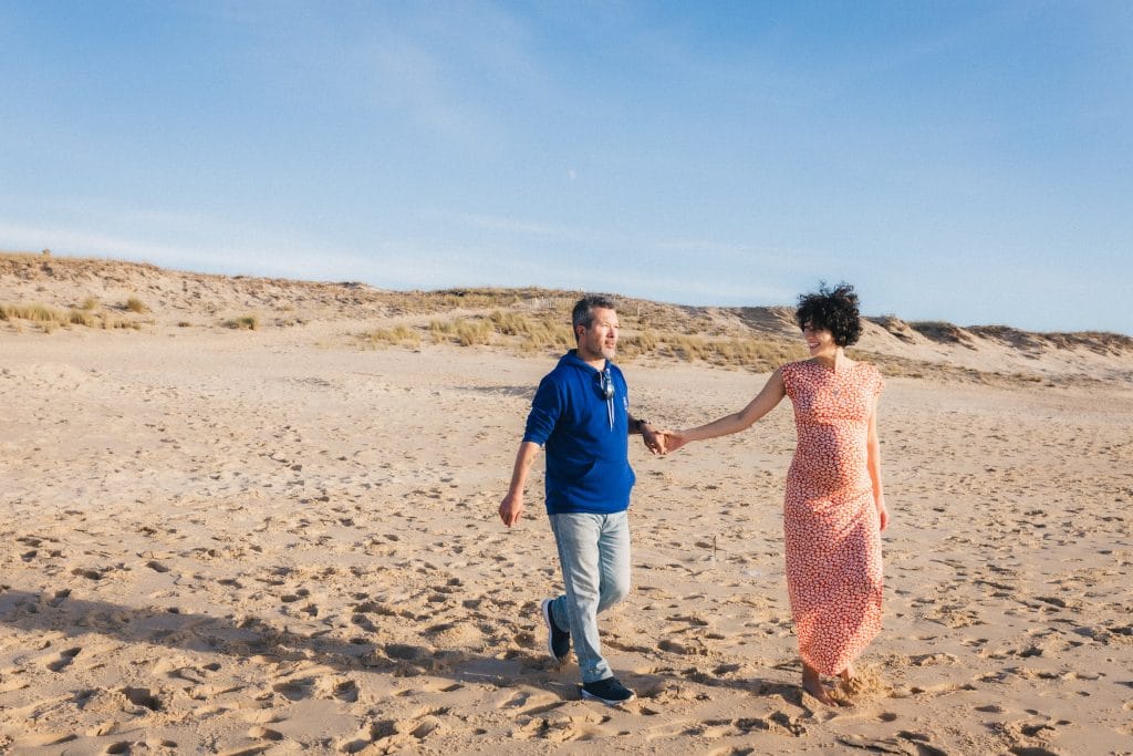 Les futurs parents marchent main dans la main sur le sable, se dirigeant vers l'océan