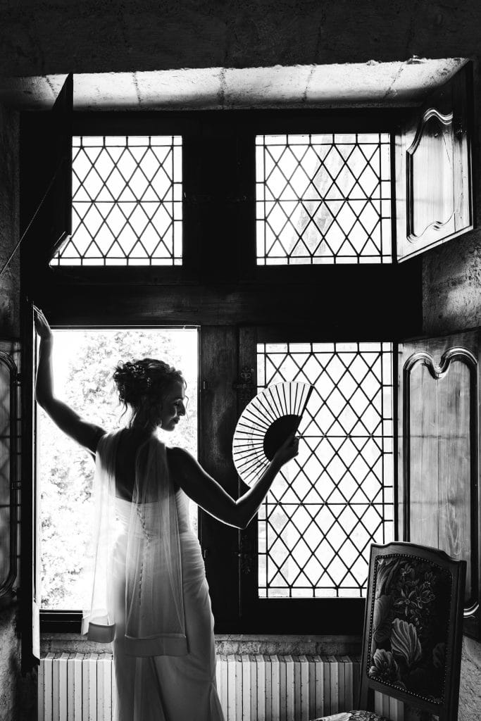 La mariée prend la pose face à la fenêtre en agitant son éventail lors de son mariage au château Sainte-Foy