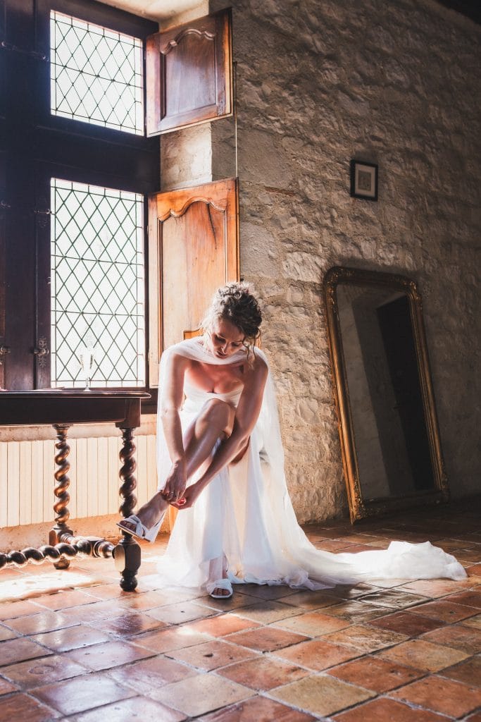 La mariée, assise sur une chaise à côté de la fenêtre, attache les brides de ses chaussures