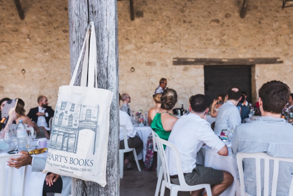 Vue extérieure de la salle de réception, avec au premier plan un tote bag suspendu à une poutre. Les invités écoutent un discours.