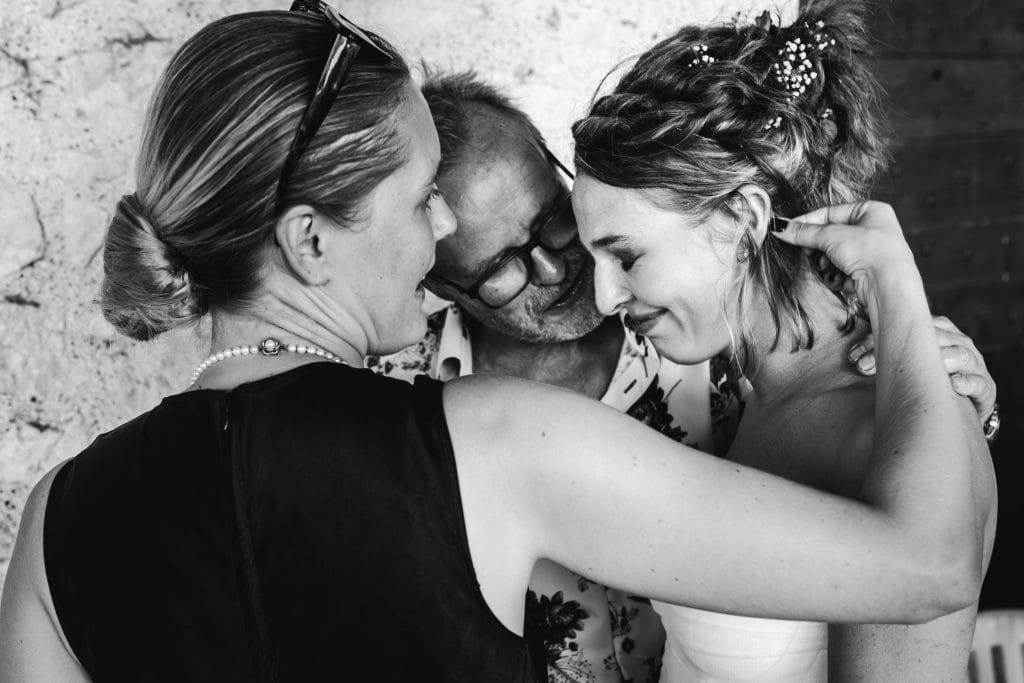 La mariée est émue, entourée de son père et de sa soeur qui lui remet une mèche de cheveux derrière l'oreille, pendant son repas de mariage au château Sainte-Foy