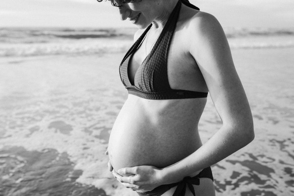 La future maman, en maillot de bain au bord de l'eau, regarde tendrement vers son ventre rond