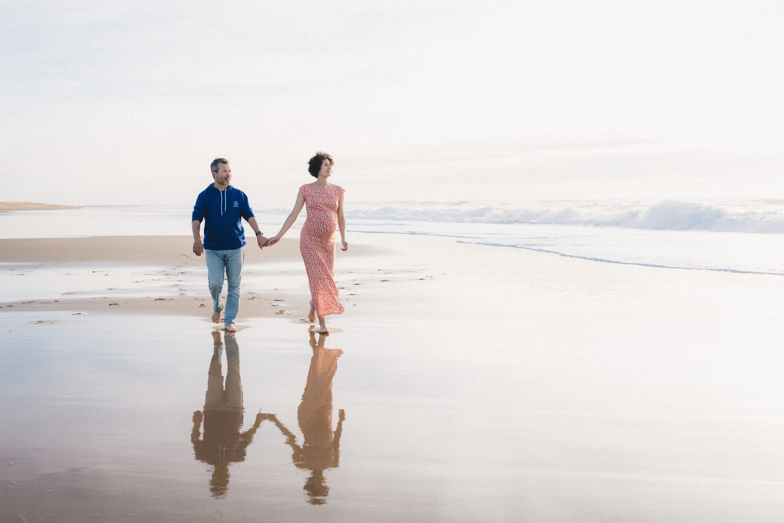 Les futurs parents se tiennent par la main et marchent le long de la plage, leurs silhouettes se reflètent dans l'eau.