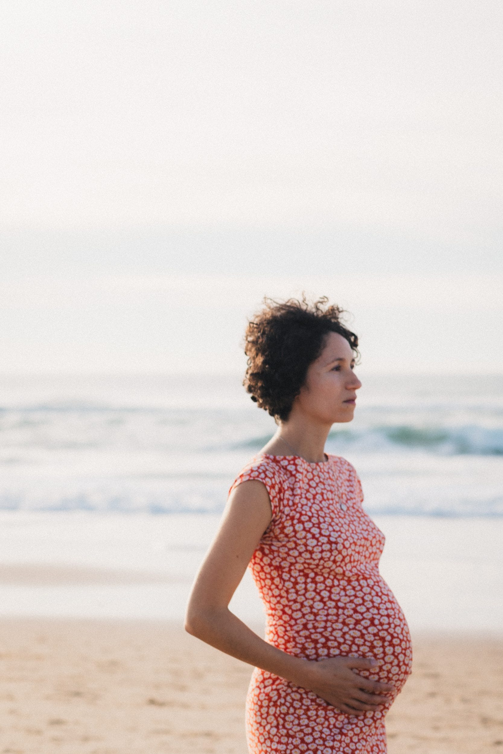 La future maman regarde au loin la plage, d'un air mélancolique, en caressant son ventre rond, lors de sa séance grossesse à Lacanau