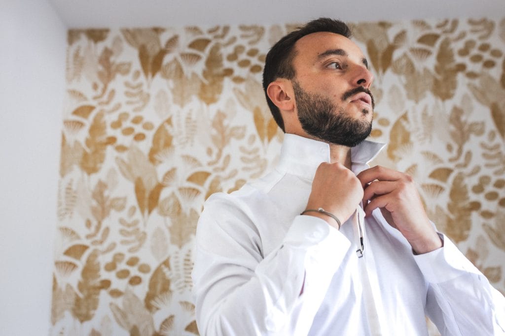 Le marié boutonne le col de sa chemise dans une chambre