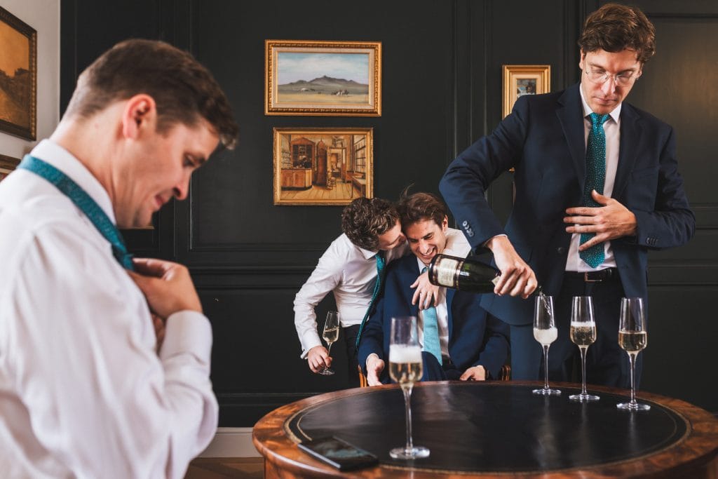 Le marié et ses témoins partagent un moment de convivialité en riant, l'un serre les coupes de champagne et l'autre essayant de nouer sa cravate