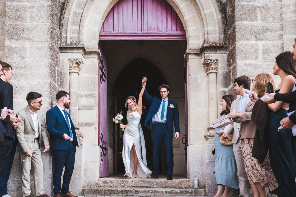Les mariés sortent triomphants de l'église, levant les bras en l'air, pour leur mariage à Saint-Emilion