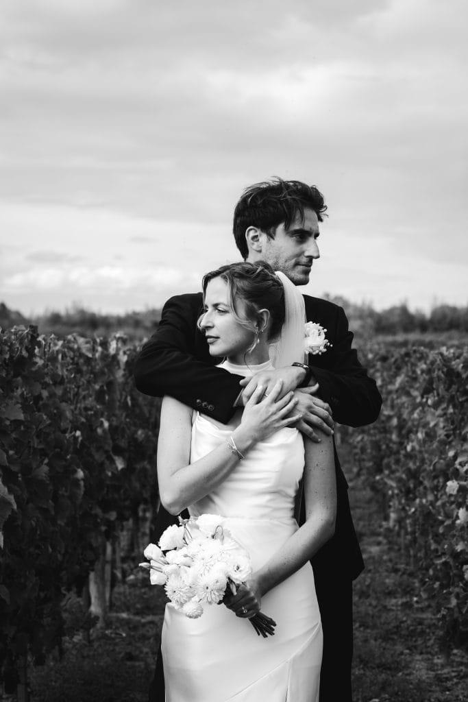 Dans les vignes lors de leur mariage à Saint-Emilion, les mariés se tiennent tendrement enlacés en regardant chacun dans une direction différente