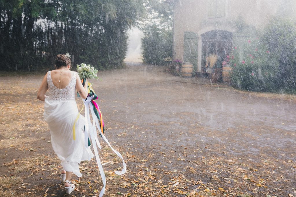 De dos, la mariée court, son bouquet à la main, sous une pluie diluvienne