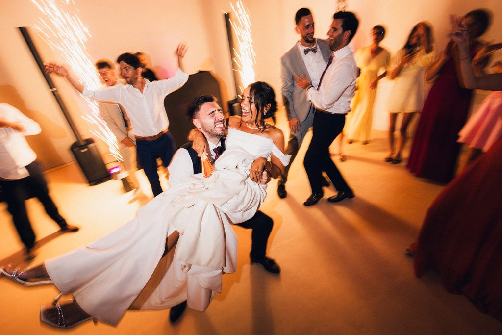 Sur le dancefloor, le marié porte dans ses bras son épouse, alors que les invités dansent et que les étincelles jaillissent du sol.