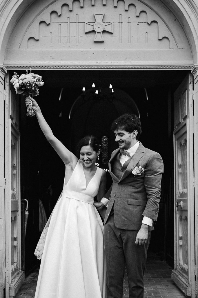 Les mariés sortent triomphants de l'église, les bras en l'air avec le bouquet dans la main